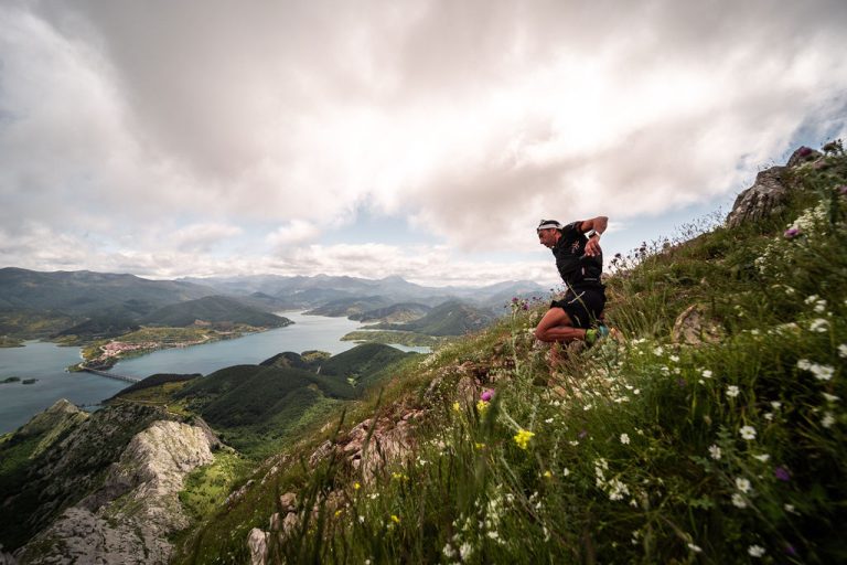 Corredor de trail running descendiendo una ladera técnica de Riaño Trail Run, con vistas panorámicas al embalse de Riaño y las montañas bajo un cielo nublado